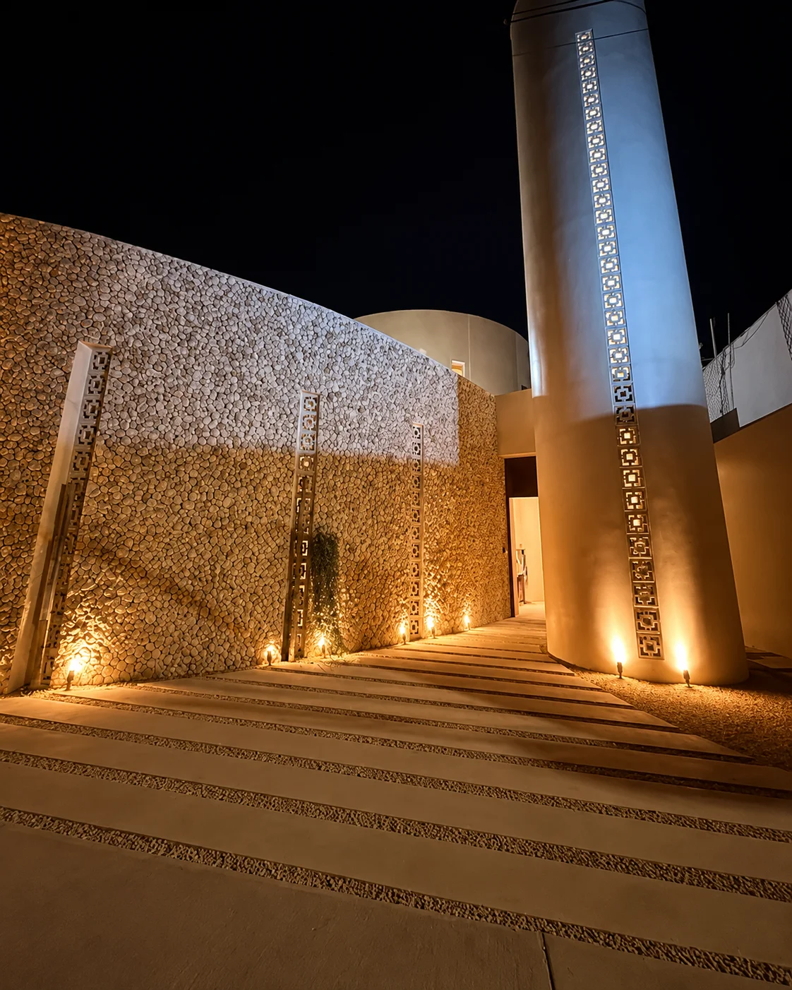 Exterior de la mezquita iluminado de noche, con el minarete y un camino de piedra